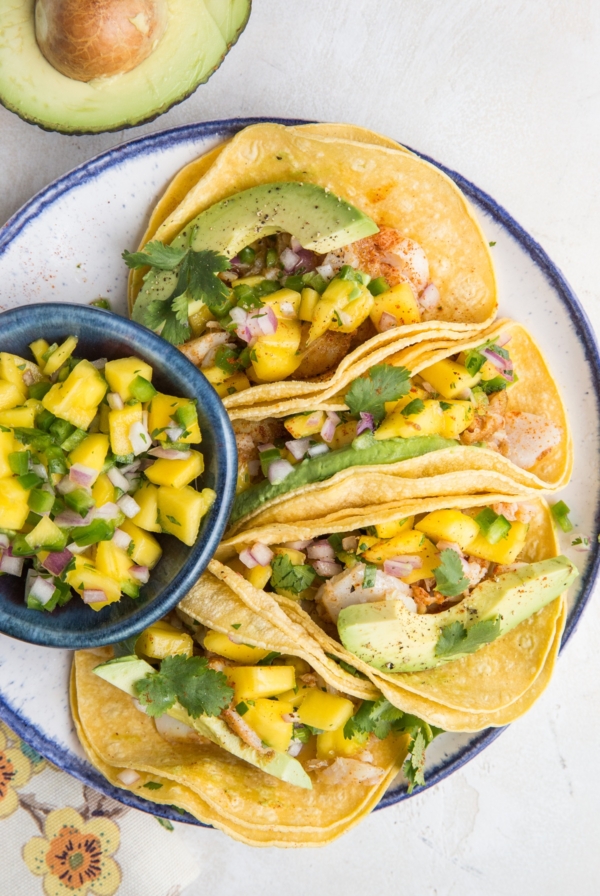 Three Fish Tacos with Mango Salsa and fresh cilantro on corn tortillas sitting on a blue-rimmed plate with a floral napkin to the side and a bowl of fresh mango salsa.
