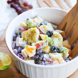 White bowl of fruit salad with a bunch of grapes in the background and a fresh basil leaf in front of the bowl.
