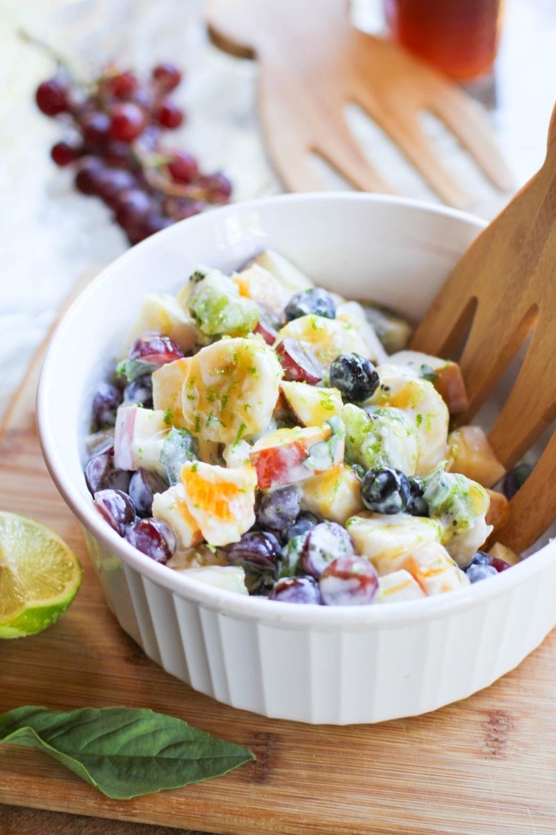 White bowl of fruit salad with a bunch of grapes in the background and a fresh basil leaf in front of the bowl.