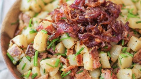 German-Style Potato Salad with bacon, caramelized onions, and chives in a bowl with the caramelized onions on top. The wooden bowl is on top of a striped napkin and the dish is ready to serve.