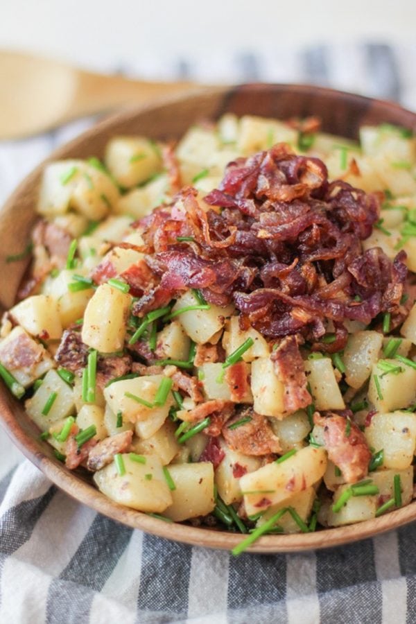 German-Style Potato Salad with bacon, caramelized onions, and chives in a bowl with the caramelized onions on top. The wooden bowl is on top of a striped napkin and the dish is ready to serve.