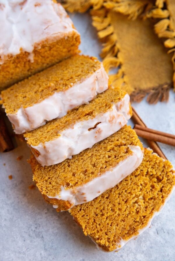 Pumpkin spice bread cut into slices with a glaze on top, cinnamon sticks and a golden napkin in the background.