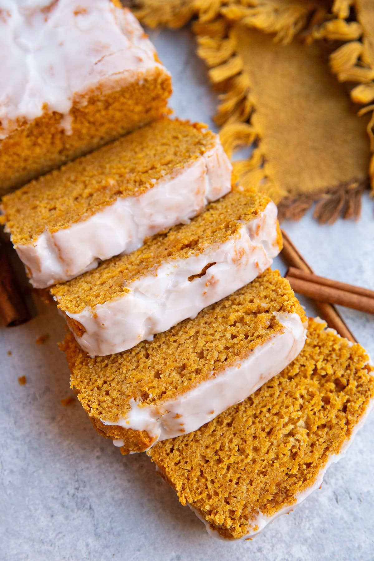 Pumpkin spice bread cut into slices with a glaze on top, cinnamon sticks and a golden napkin in the background.
