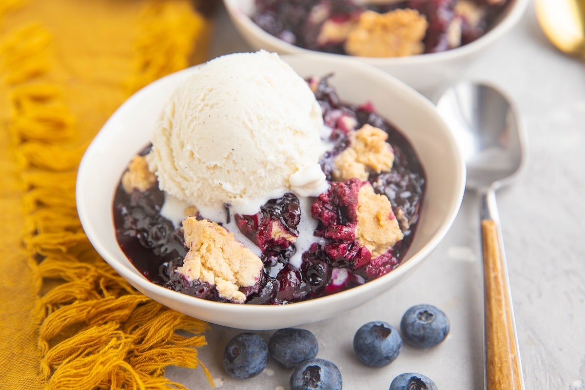 Two white bowls full of blueberry crumble with a scoop of vanilla ice cream on top. Wood inlay spoon to the side and fresh blueberries.