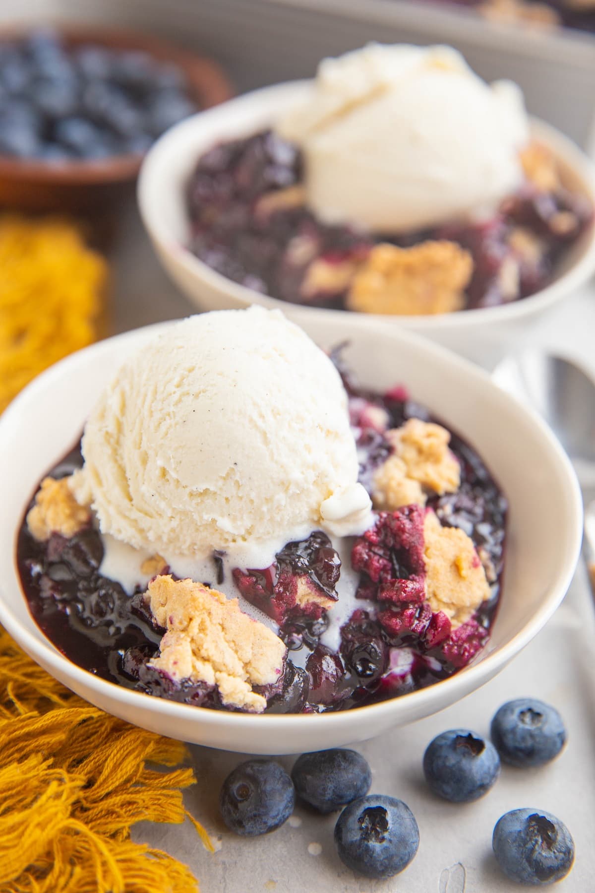 Two white bowls of blueberry cobbler with scoops of ice cream on top.