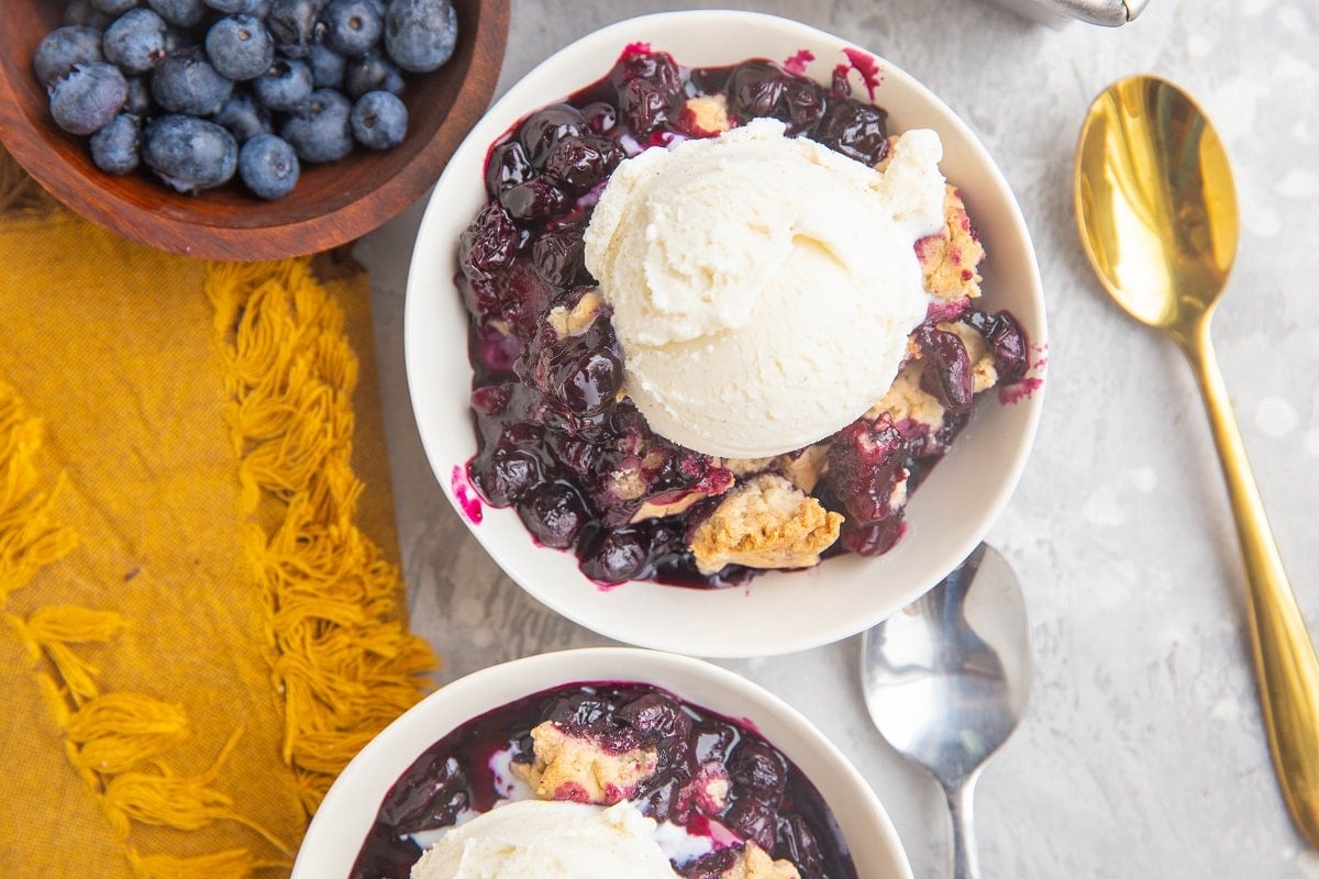 Two bowls of blueberry cobbler with spoons and a bowl of fresh blueberries to the side.