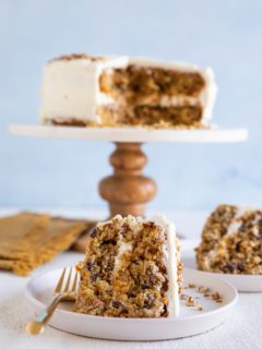 Two plates with slices of carrot cake on top and a cake stand in the background with the rest of the cake.