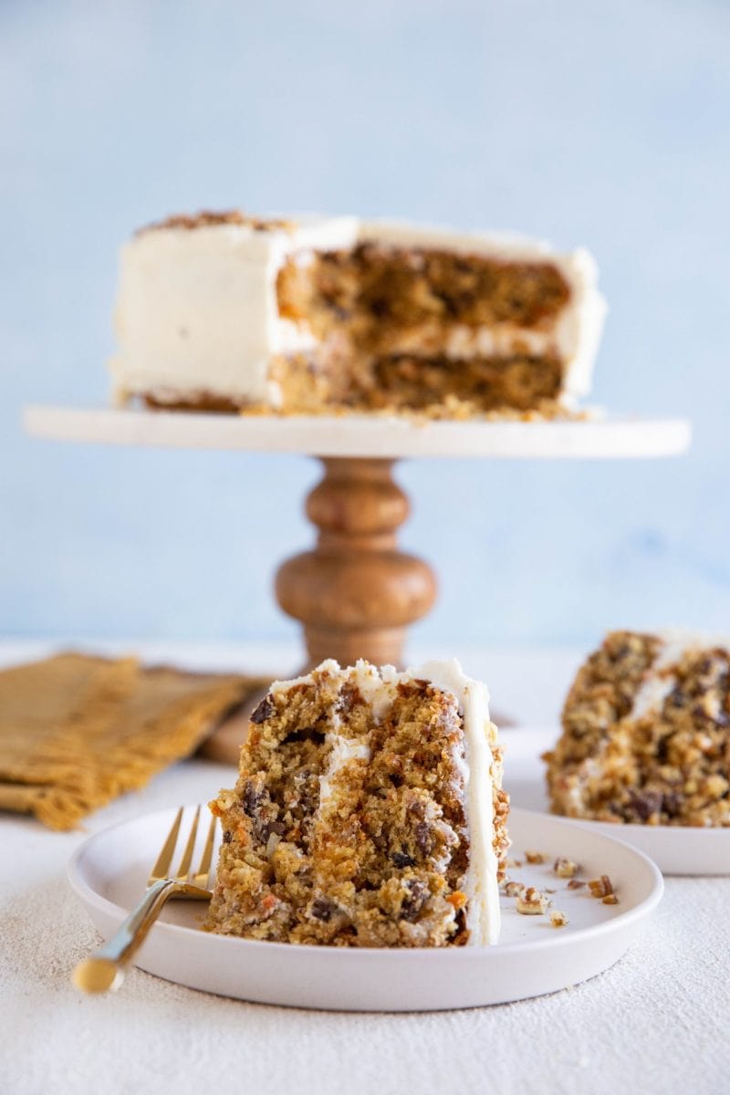 Two plates with slices of carrot cake on top and a cake stand in the background with the rest of the cake.