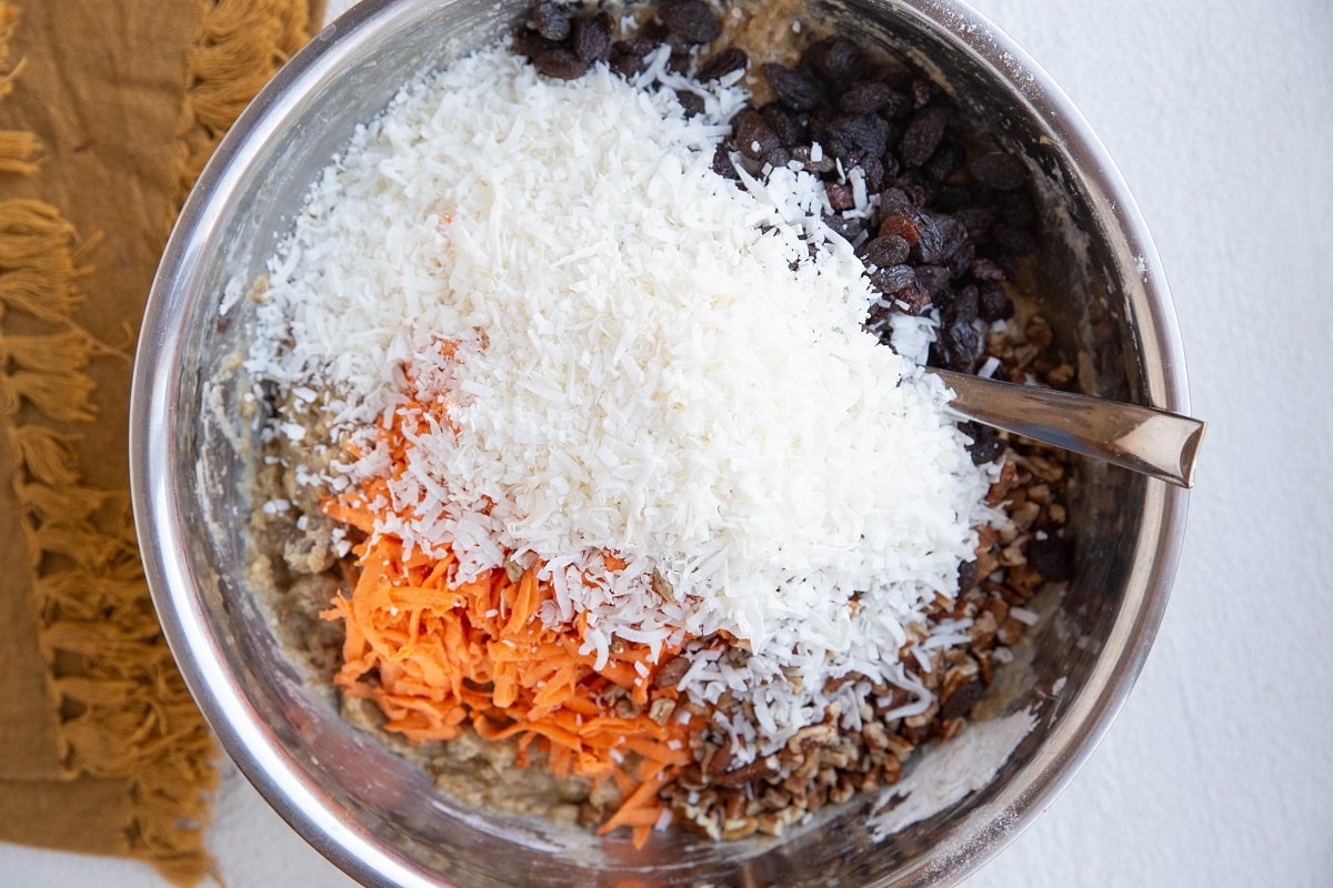 Carrot cake batter with shredded coconut, raisins, pecans, and grated carrot on top, ready to be mixed in.