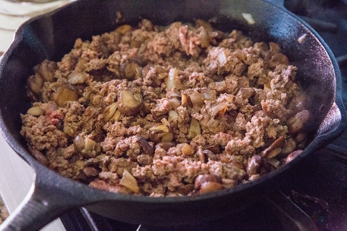 ground beef, mushrooms, and onions cooking in a cast iron skillet.