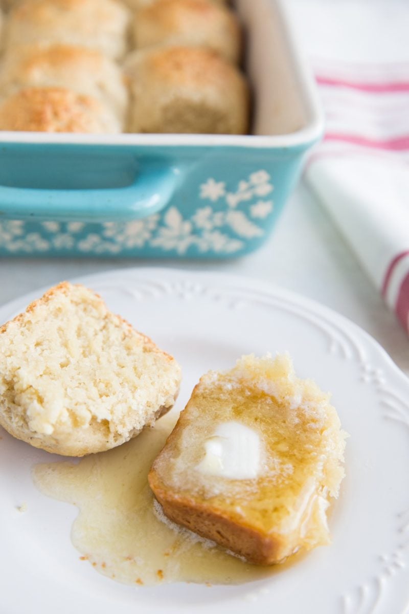 Dairy-Free Gluten-Free Hawaiian Roll on a plate sliced in half and slathered with melted butter and honey with the dish of the rest of the rolls in the background.