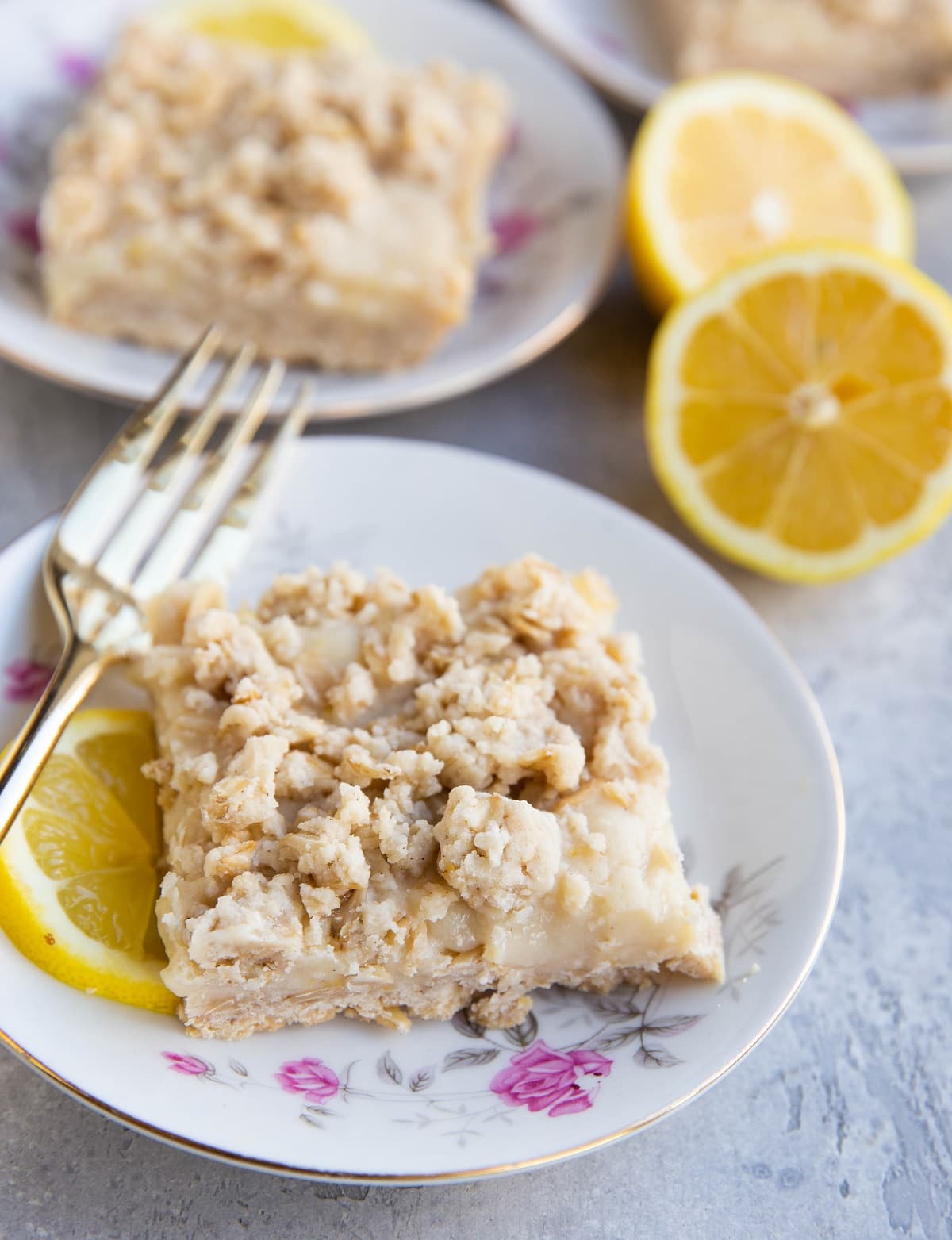 Three decorative plates with roses on them with a lemon crumb bar on top and fork to the side. Fresh lemons in the background.