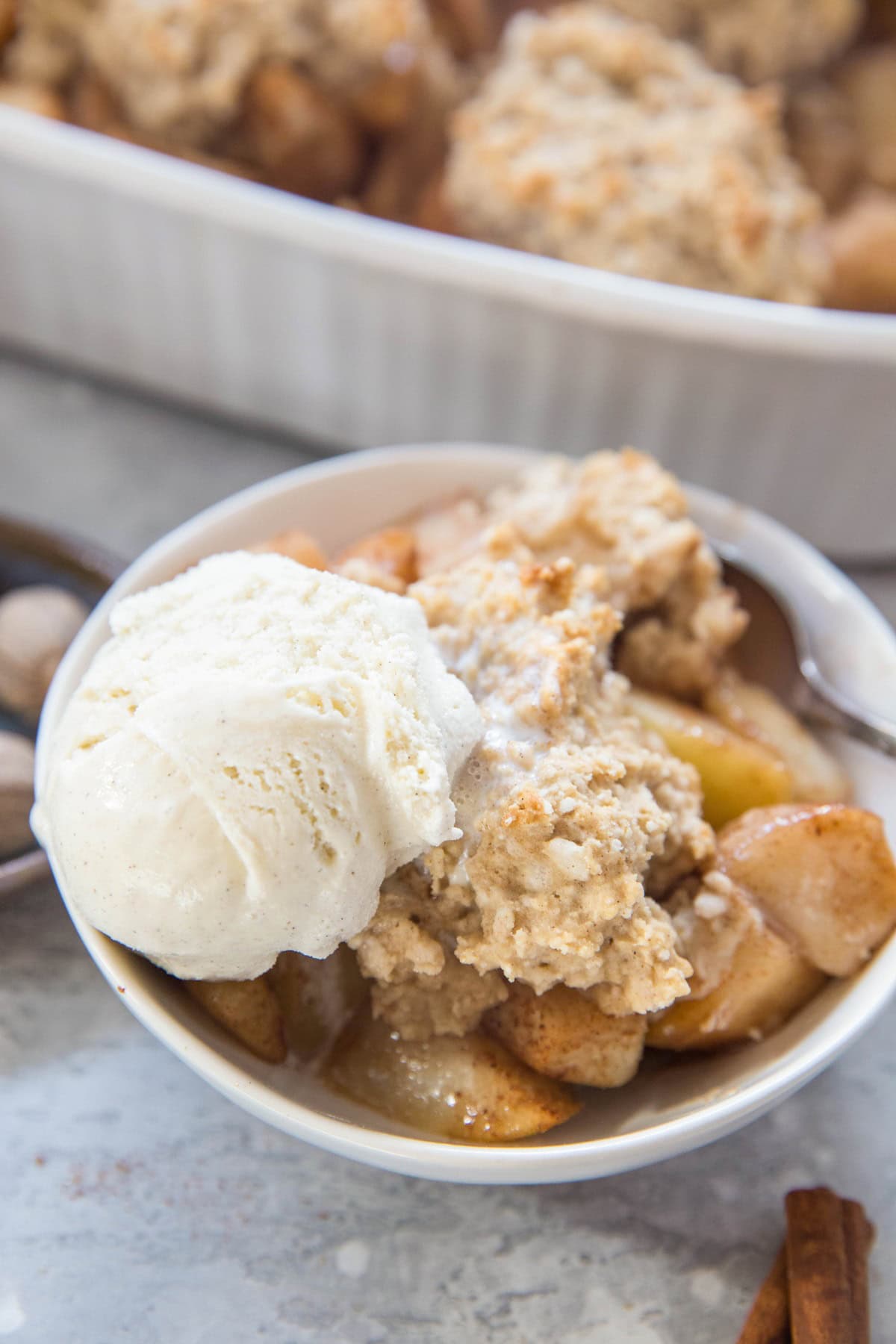 A bowl of gluten-free pear cobbler topped with vanilla ice cream sits in front of a fresh pear, warm spices, and a baking dish filled with more delicious pear crisp.