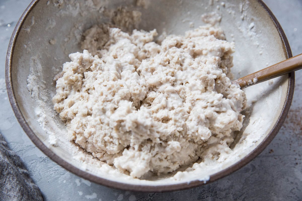 A large bowl filled with shaggy, partially mixed gluten-free pear cobbler dough. A wooden spoon rests inside the bowl, and flour is visible on the sides and around the mixture.