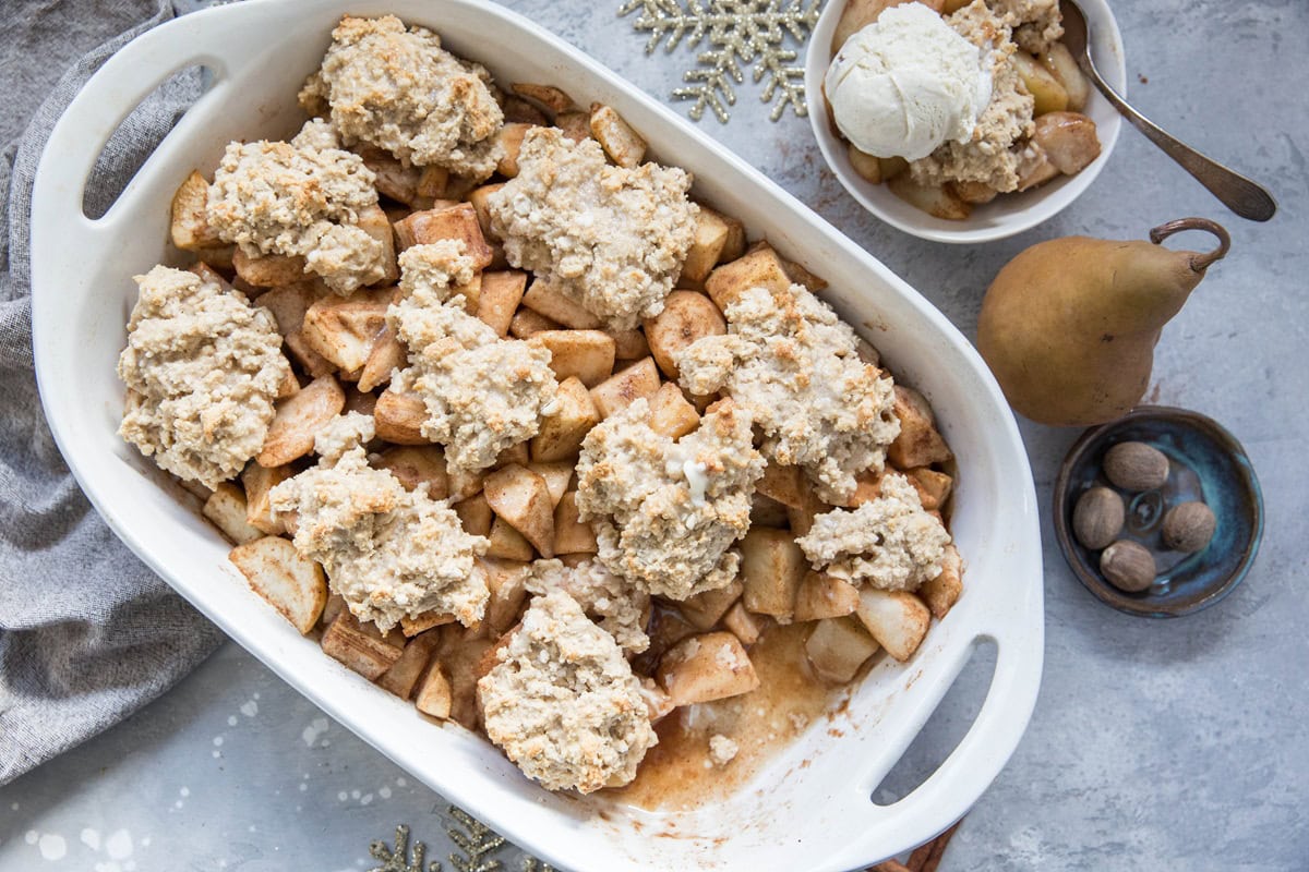 A white baking dish filled with gluten-free pear cobbler and spiced apples, topped with biscuit dough. Nearby is a bowl of cobbler with vanilla ice cream, a whole pear, whole nutmegs, and a decorative snowflake.