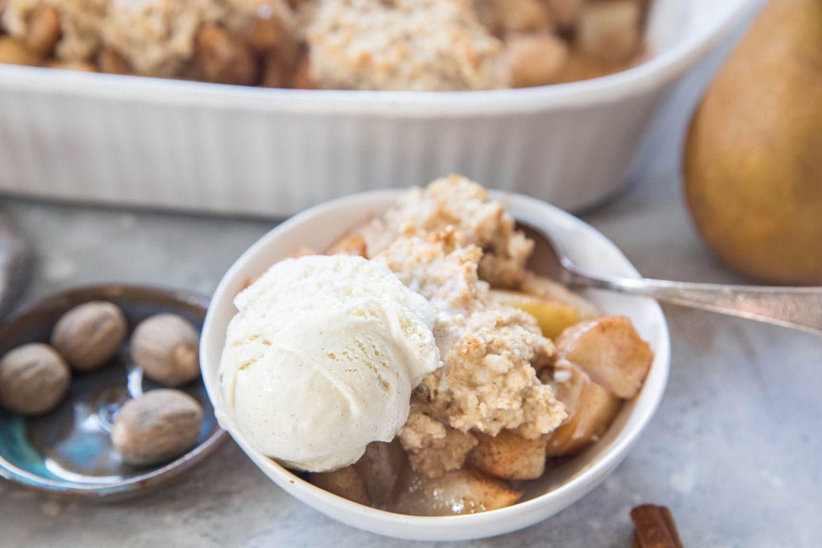 A bowl of apple crumble topped with vanilla ice cream, a spoon inside. Nearby, part of a baking dish with more crumble and a gray napkin are visible, evoking the cozy charm of a homemade gluten-free pear cobbler.
