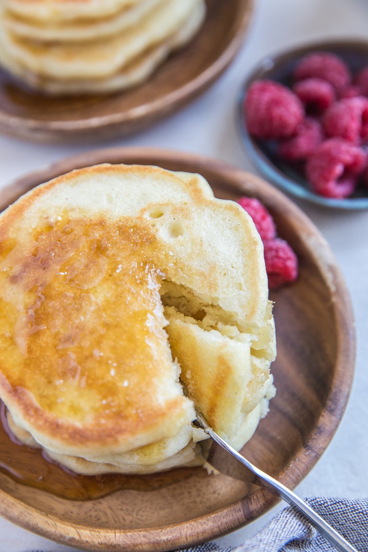 Wooden plate with a stack fluffy pancakes with a fork cutting a bite out of the stack. Fresh raspberries in the background.