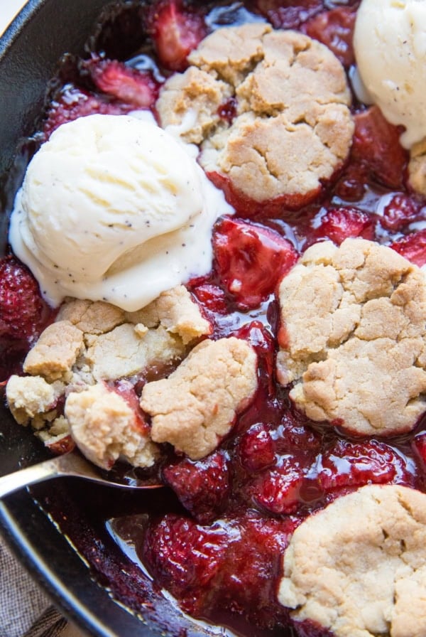 Strawberry cobbler in a large cast iron skillet with scoops of melted ice cream on top and a spoon scooping out some of the cobbler.