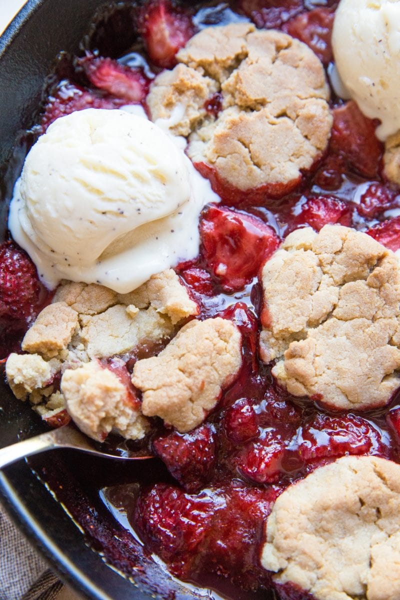 Strawberry cobbler in a large cast iron skillet with scoops of melted ice cream on top and a spoon scooping out some of the cobbler.