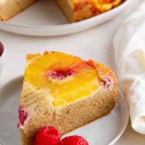 Slice of healthy pineapple upside down cake on a white plate with fresh raspberries to the side