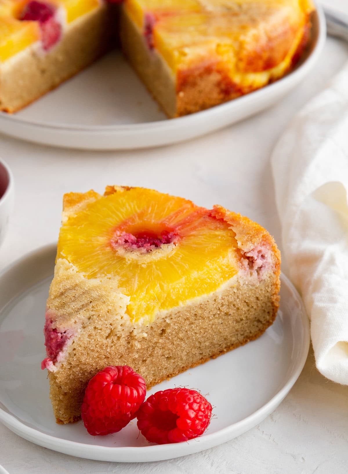 Slice of healthy pineapple upside down cake on a white plate with fresh raspberries to the side