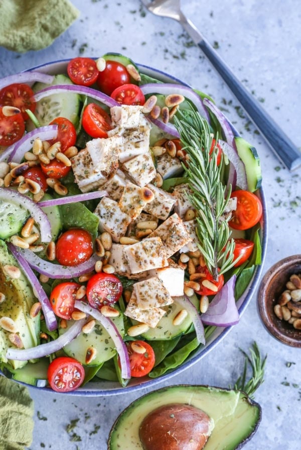 Herby Greek Spinach Salad in a large bowl with a sliced open avocado, dried herbs, and a napkin to the side.