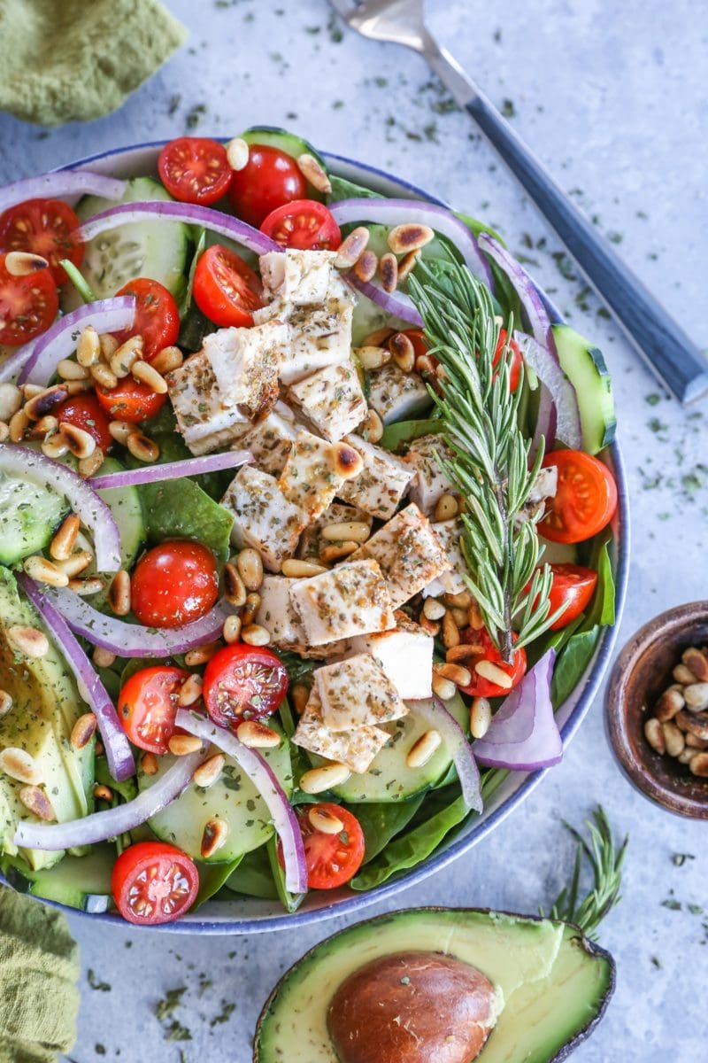Herby Greek Spinach Salad in a large bowl with a sliced open avocado, dried herbs, and a napkin to the side.