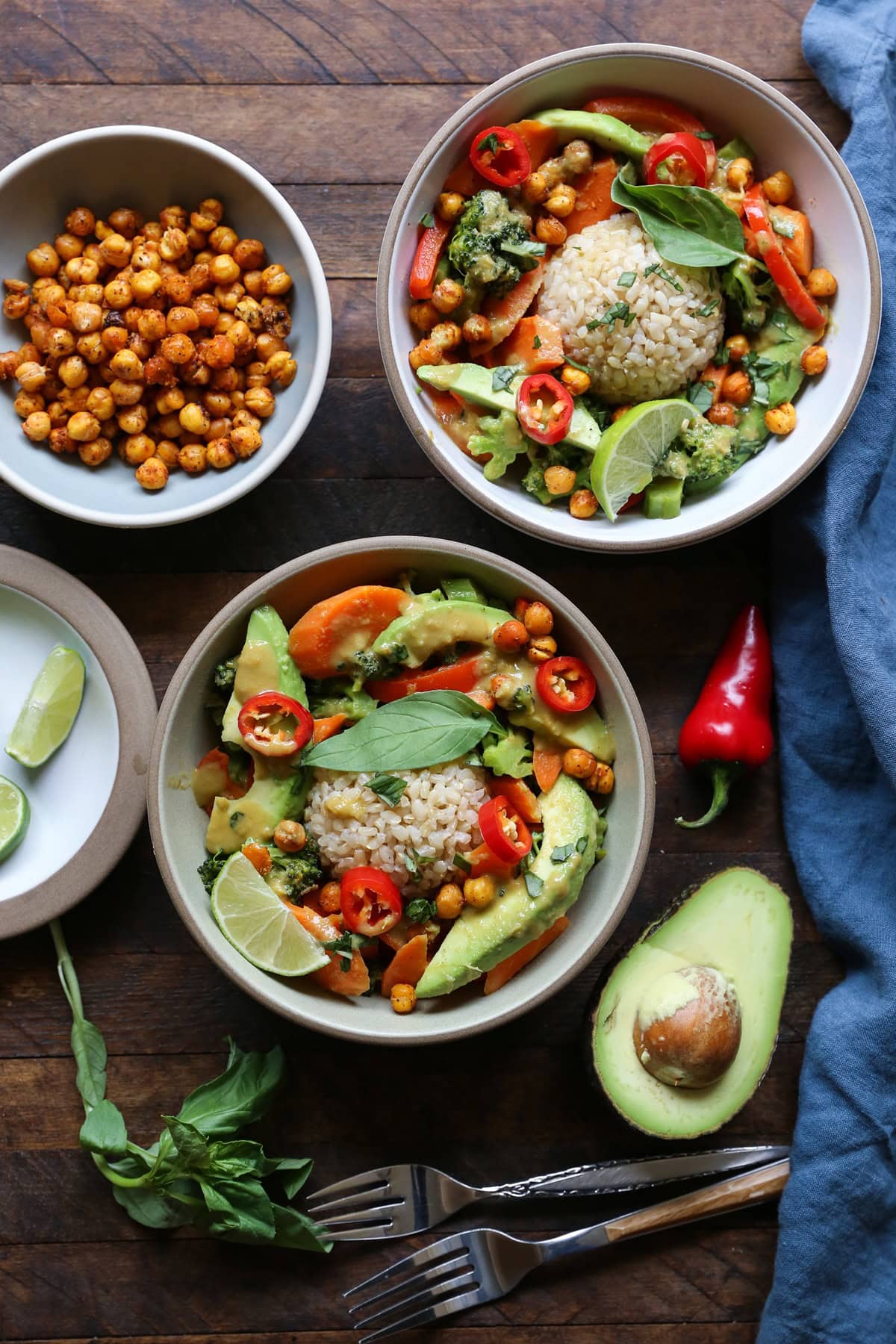 Two ceramic bowls full of curry vegetables and avocado with fresh avocado, basil, and forks to the side on a wooden table.
