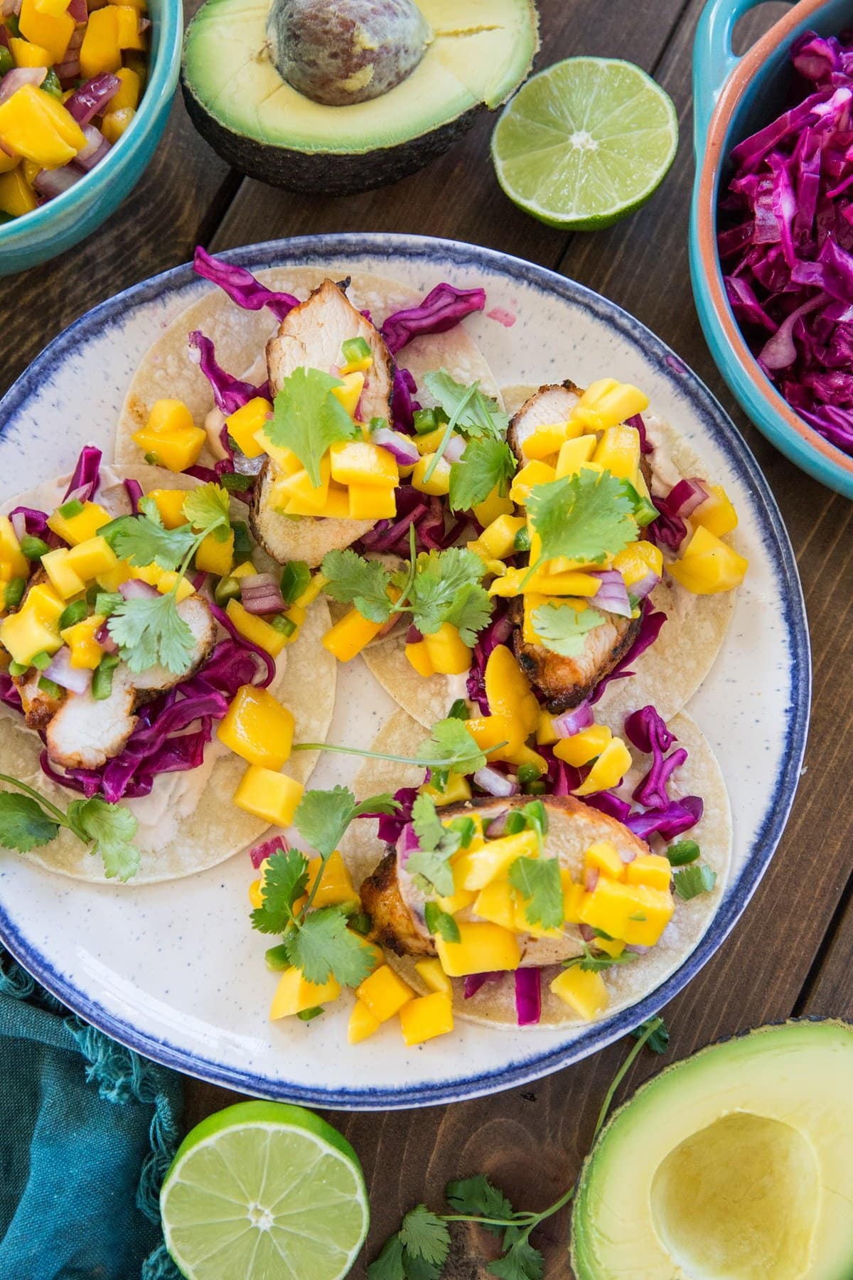 Plate of three grilled chicken tacos topped with mango salsa with fresh lime and avocado to the side.