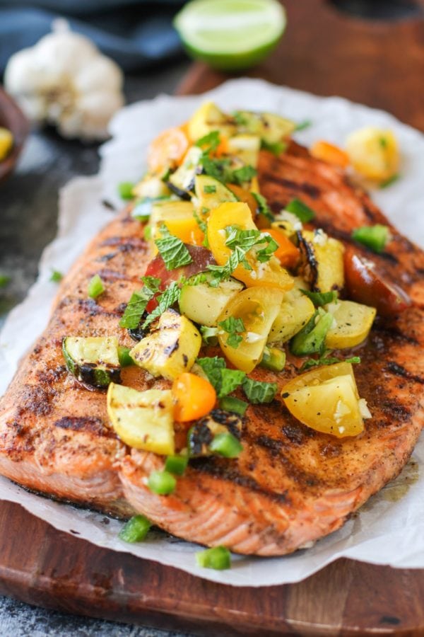 large piece of grilled salmon on a piece of parchment paper on a cutting board with grilled zucchini, tomatoes, green onions and fresh herbs on top. Ready to serve.
