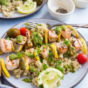 Two plates of salmon skewers with lemon slices, pesto sauce, and quinoa. A napkin and a glass of white wine to the side.