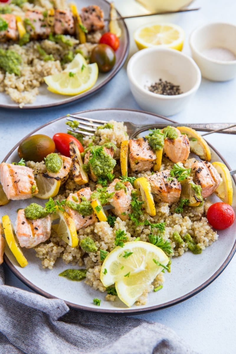 Two plates of salmon skewers with lemon slices, pesto sauce, and quinoa. A napkin and a glass of white wine to the side.