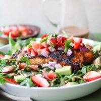 White bowl with grilled salmon on top of salad with strawberry salsa and homemade balsamic vinaigrette with a napkin to the side and balsamic vinaigrette in the background.