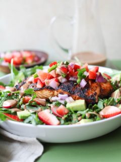 White bowl with grilled salmon on top of salad with strawberry salsa and homemade balsamic vinaigrette with a napkin to the side and balsamic vinaigrette in the background.