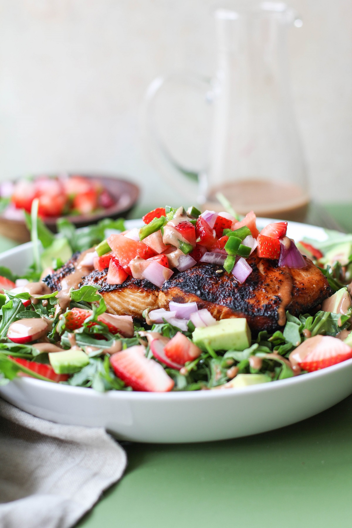 White bowl with grilled salmon on top of salad with strawberry salsa and homemade balsamic vinaigrette with a napkin to the side and balsamic vinaigrette in the background.