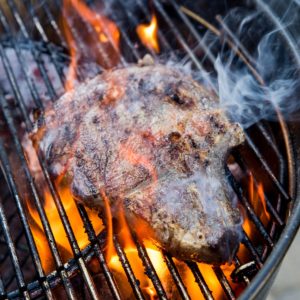 Grilling steak on a charcoal grill with a large flare up, flames engulfing the steak.