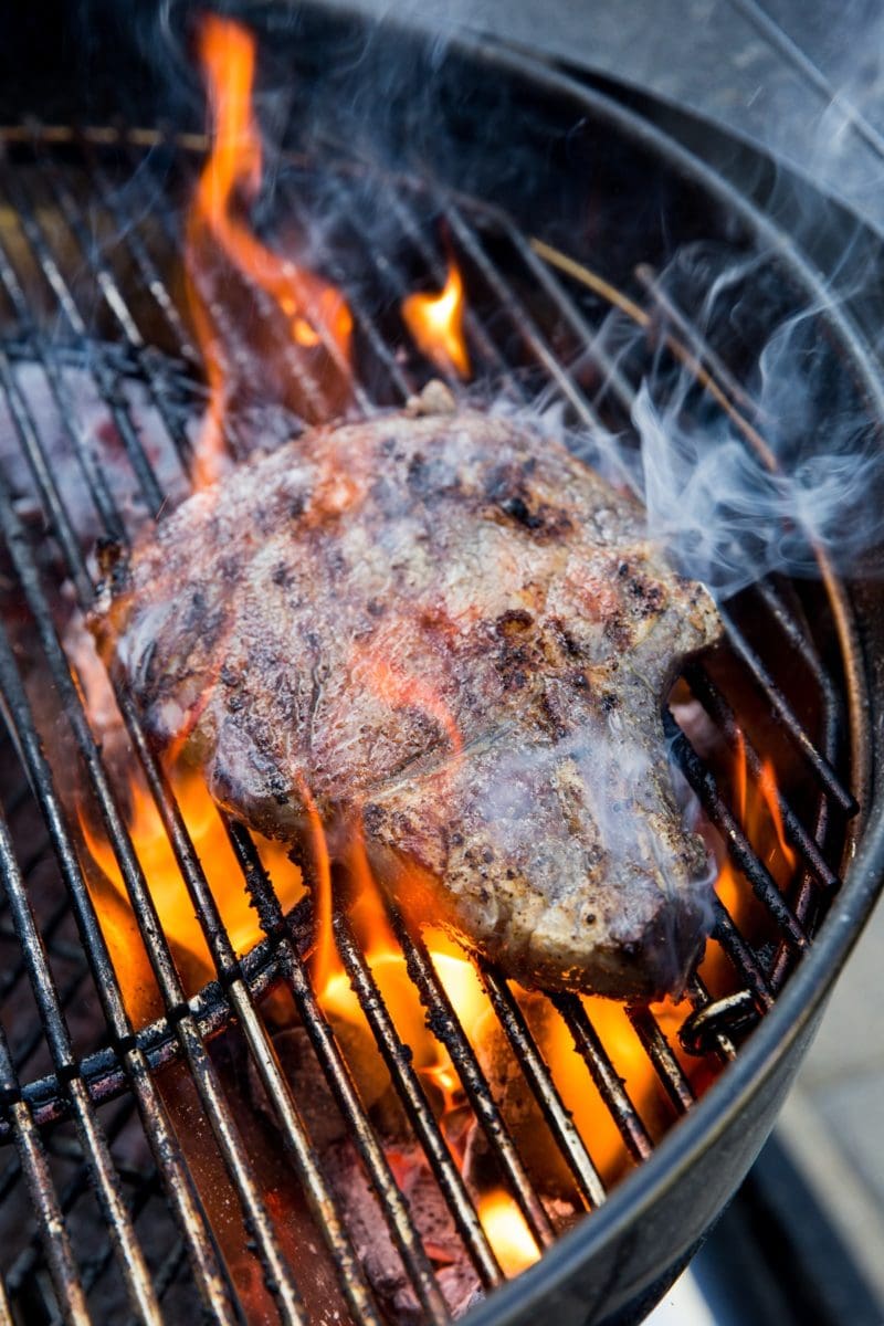 Grilling steak on a charcoal grill with a large flare up, flames engulfing the steak.
