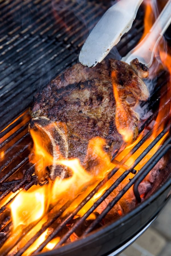 Steak being flipped by metal tongs on a charcoal grill with a large fire flare up.