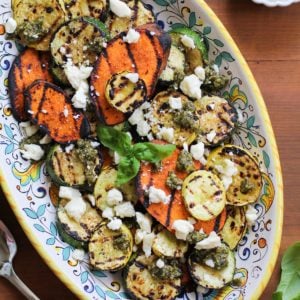 Platter of grilled sweet potatoes, yellow squash, and zucchini with feta cheese sprinkled on top and pesto sauce drizzled over everything.