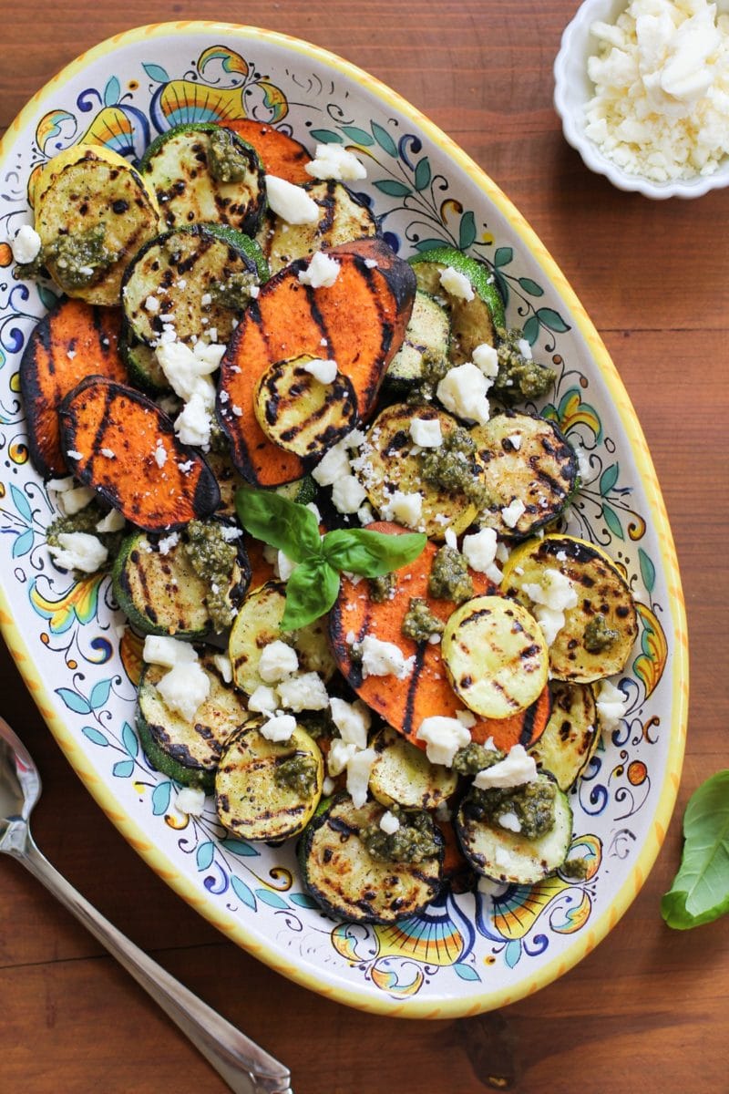 Platter of grilled sweet potatoes, yellow squash, and zucchini with feta cheese sprinkled on top and pesto sauce drizzled over everything.