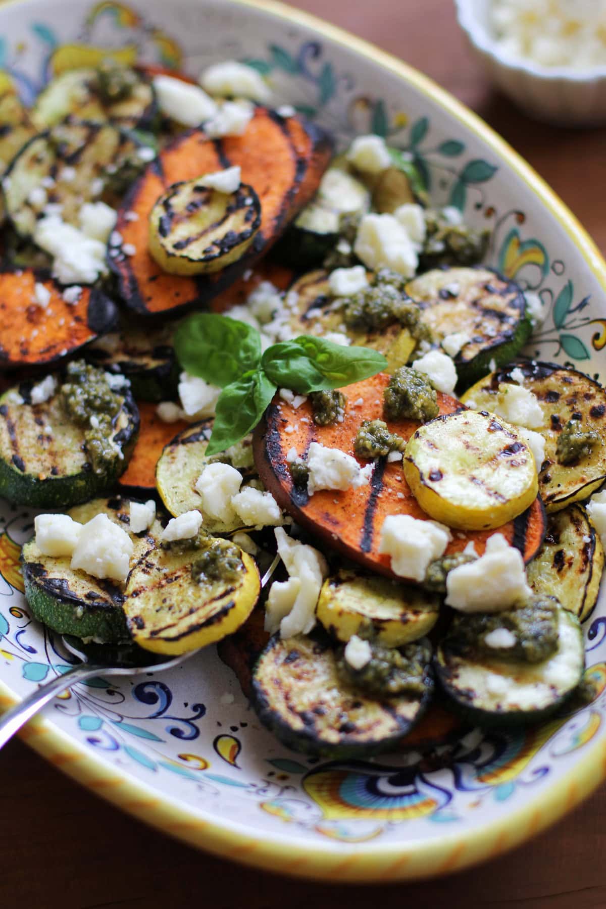 Large serving plate with grilled vegetables on top, sprinkled with feta cheese and drizzled with pesto sauce.