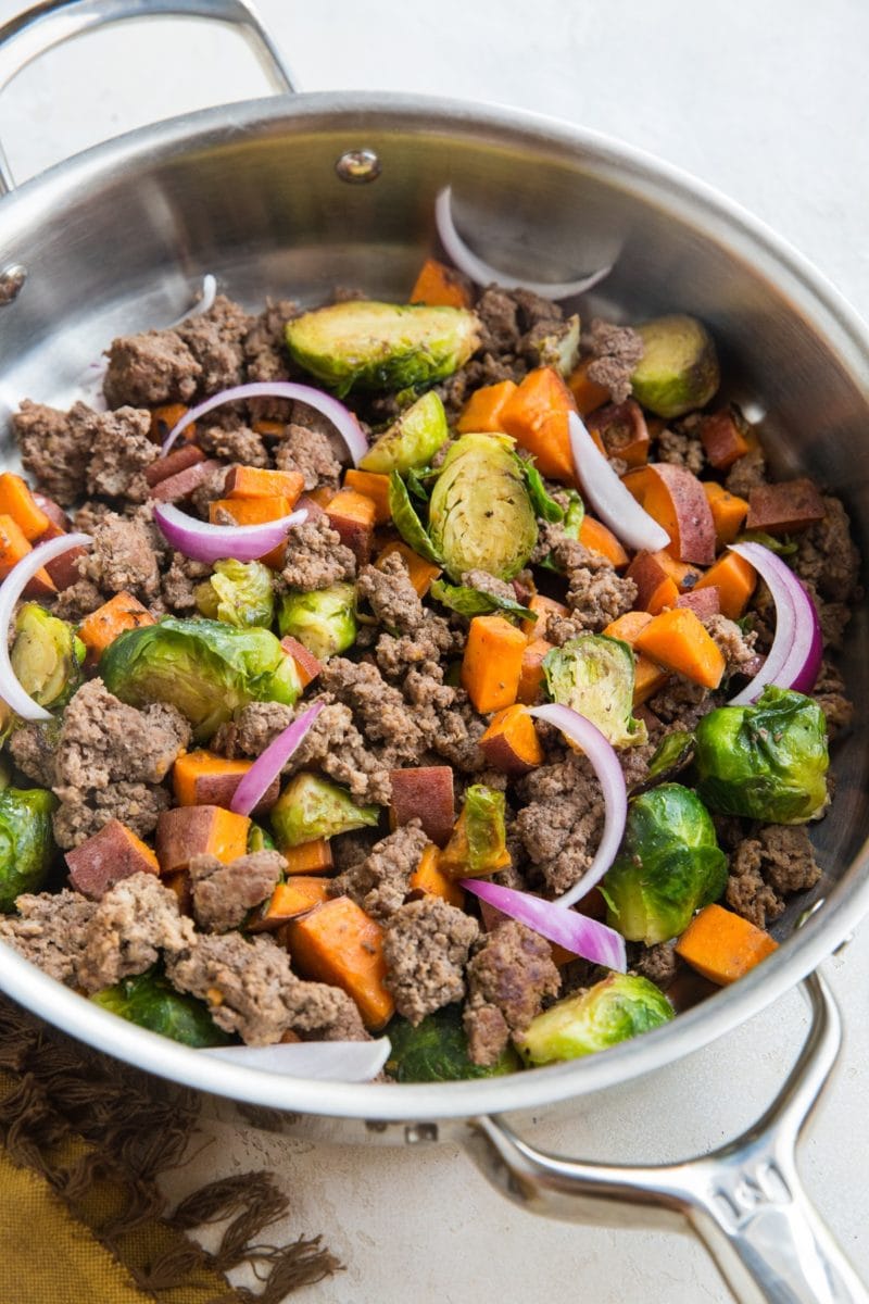 large stainless steel skillet with cooked ground beef, sweet potatoes, and brussel sprouts.