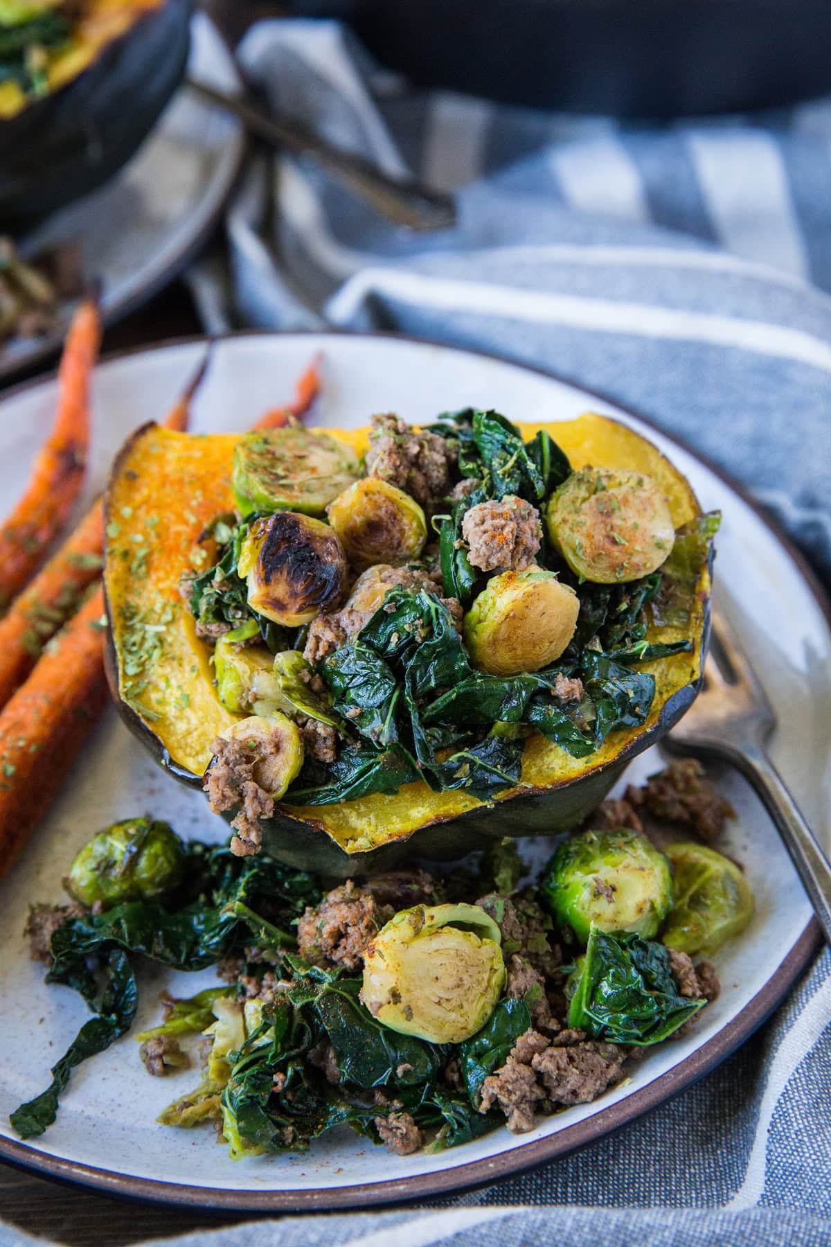 Half of an acorn squash stuffed with ground beef, kale, and brussels sprouts on a plate with roasted carrots to the side.