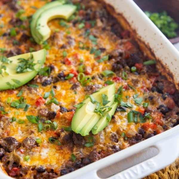 Casserole dish with ground beef casserole inside. Fresh avocado and a bowl of green onions to the side.