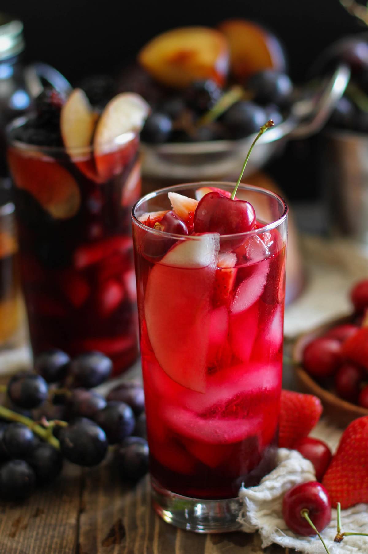 Two tall glasses of cocktails with fruit inside for a Halloween sangria recipe. The one in front is bright blood red and the one in the background is dark.