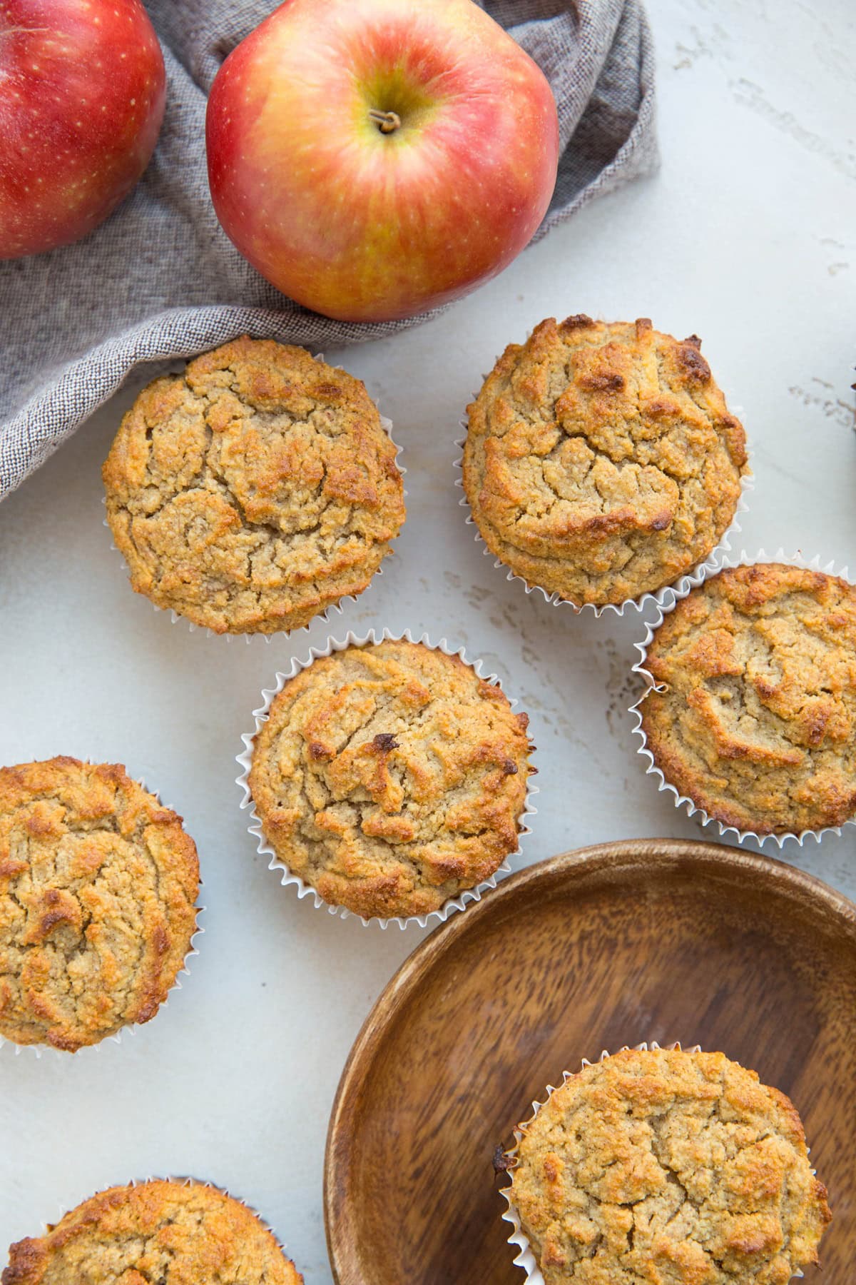 Healthy Apple Muffins on a white backdrop with fresh apples to the side and one muffin on a wooden plate. Ready to eat.