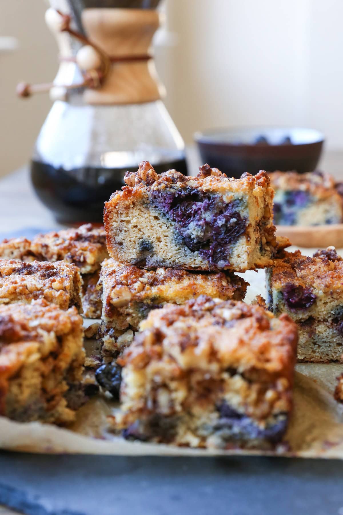 stack of paleo blueberry coffee cake with a slice full of blueberries to see the inside. A Chemex (coffee pour over) in the background for coffee.