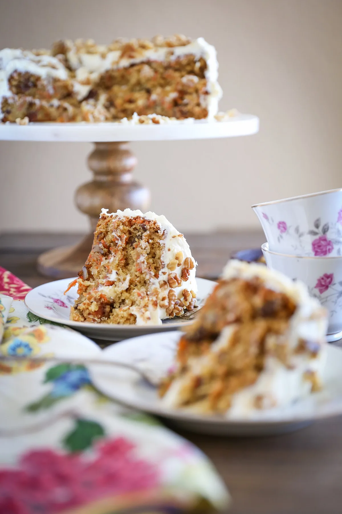 two slices of carrot cake on two plates with a cake stand with the rest of the carrot cake in the background.