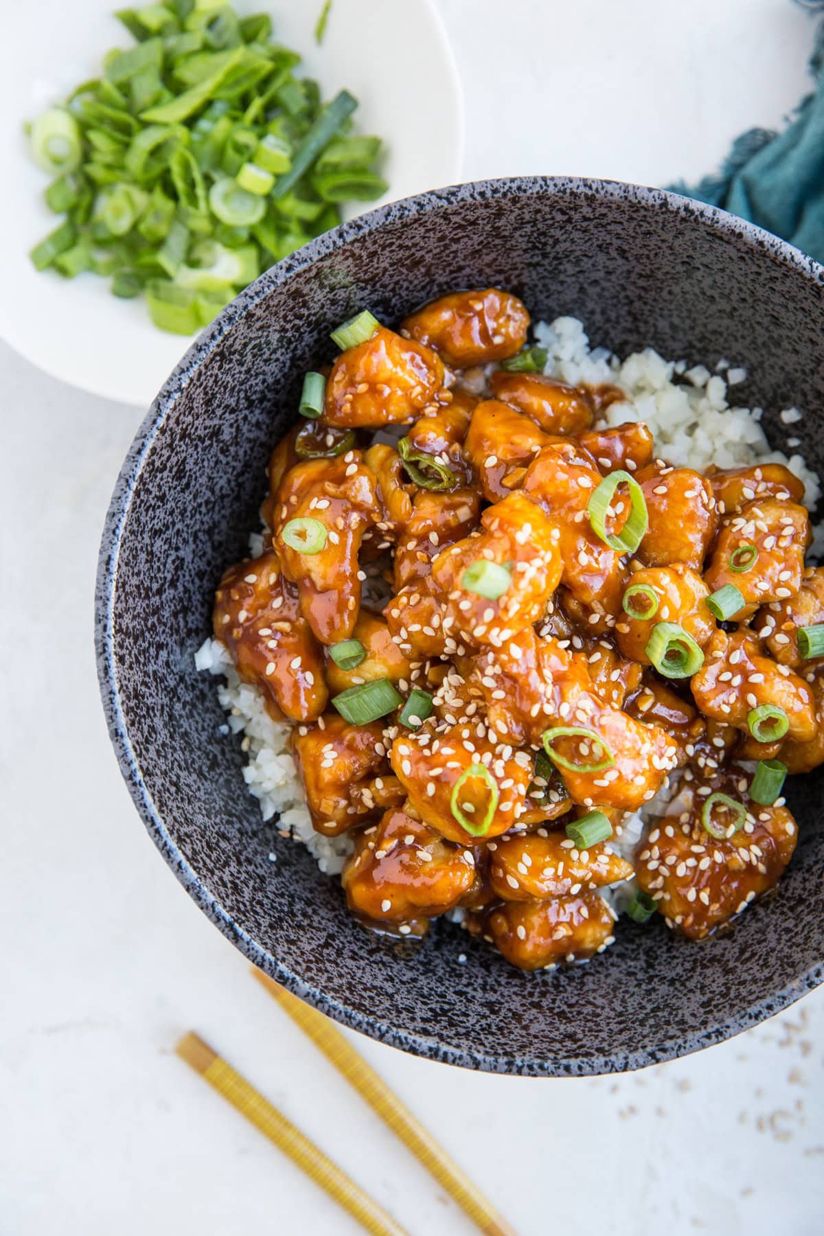 Bowl full of general chicken over rice with chopsticks to the side and a bowl of chopped green onions
