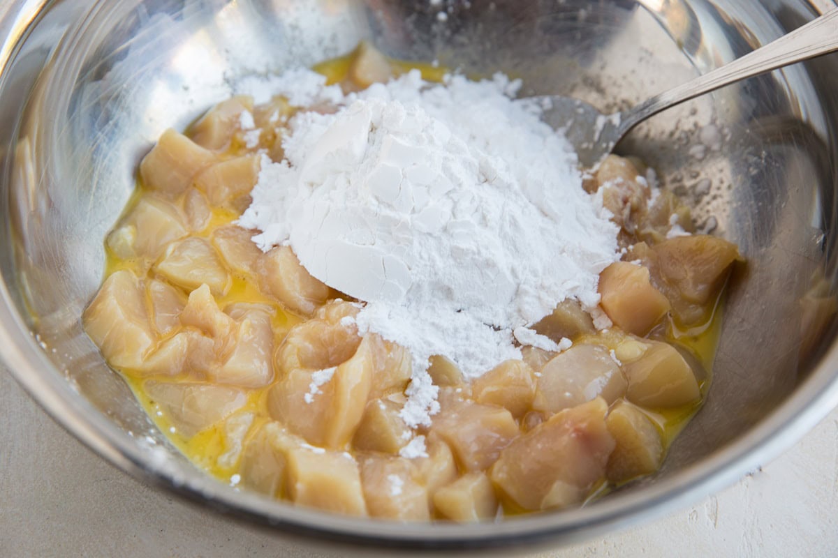 Stainless steel bowl full of chopped chicken breasts in beaten eggs with flour sprinkled on top, ready to be mixed in.